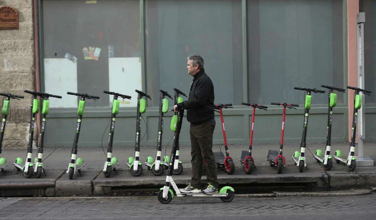 A scooter user rides past a row of scooters on Commerce Street on Tuesday, Dec 4, 2018. Scooters have rapidly made their presence felt in downtown San Antonio. For many users, the vehicle is a novelty and a handy method of transportation. But with the climbing growth of users, rules for their usage have been sketchy or unclear. Some users have gone as far as using them along the RiverWalk which is prohibited by the city. (Kin Man Hui/San Antonio Express-News)