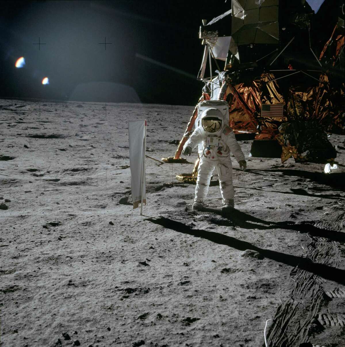 Astronaut Edwin “Buzz” Aldrin, Lunar Module pilot, stands beside the Solar-Wind Composition Experiment, facing the camera in this photo from 1969. The LM is visible behind Aldrin. Linear trails (lines) in the right foreground were formed by the cable of the surface television camera.