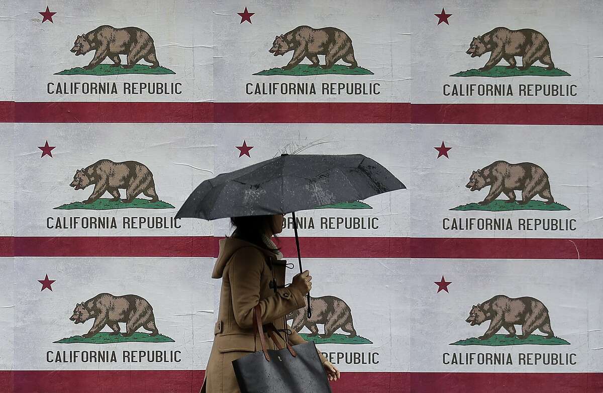 A woman carries an umbrella in the rain while walking past a billboard displaying California flags in San Francisco, Friday, Feb. 8, 2019. (AP Photo/Jeff Chiu)