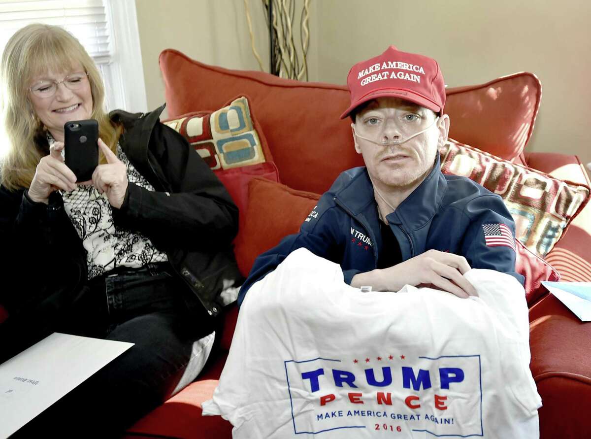 West Haven, Connecticut - Saturday, March 9, 2019: Jay W. Barrett, 44, of West Haven, terminally ill with cystic fibrosis who is a President Donald Trump fan and received a phone call from the President Tuesday right,, with visiting West Haven Mayor Nancy R. Rossi, left, shows of an autographed Make America Great Again hat, an official Donald Trump Presidential campaign jacket and a t-shirt that was among the many gifts he received from the President and his son Eric Trump Saturday afternoon delivered to his home by HUD official and personal Trump family friend Lynne Patton, who is from the New Haven area.