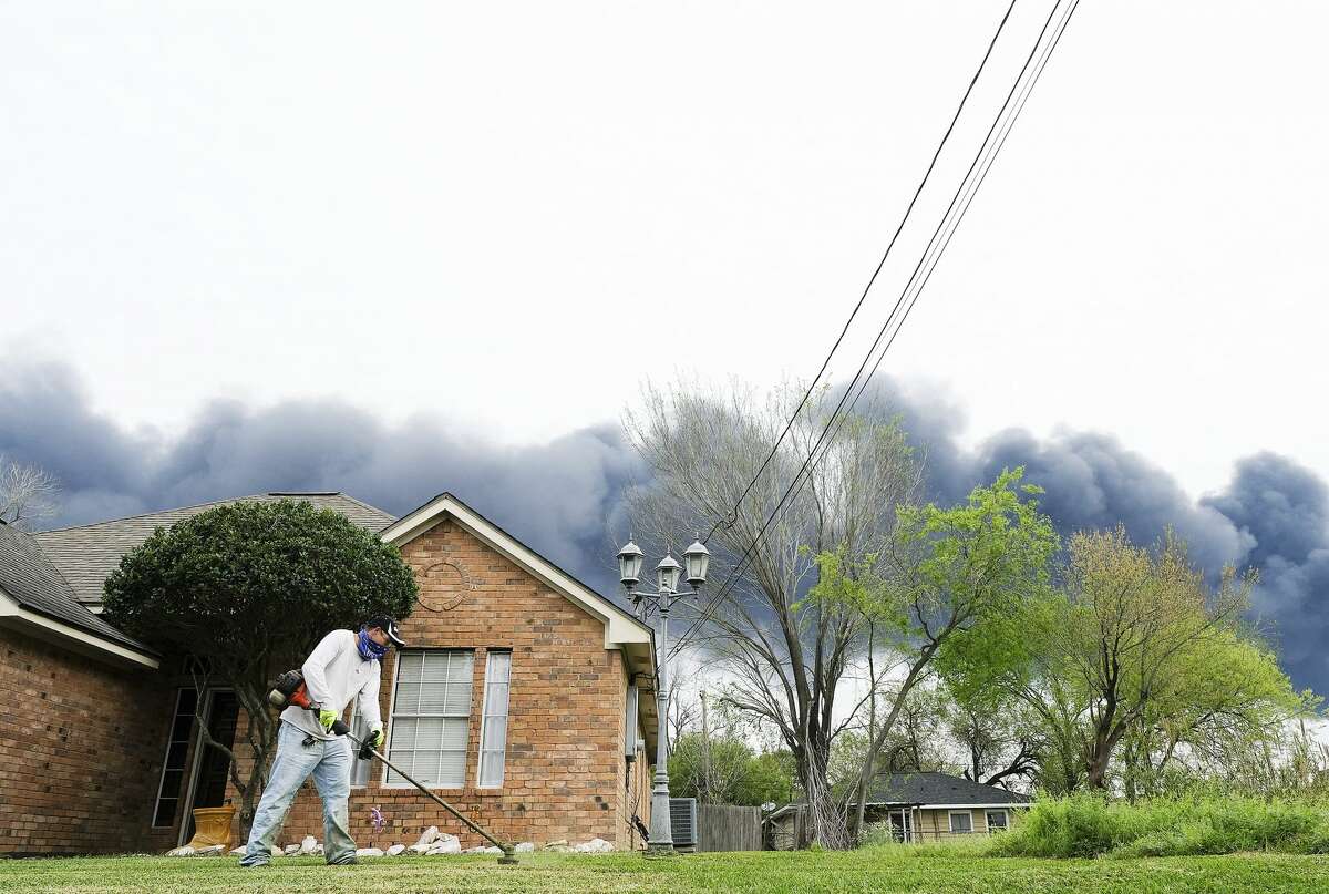 Landscaper Walter Torres works on a lawn in Deer Park while a plume from a petrochemical fire at Intercontinental Terminals Company crosses the sky on Monday, March 18, 2019.