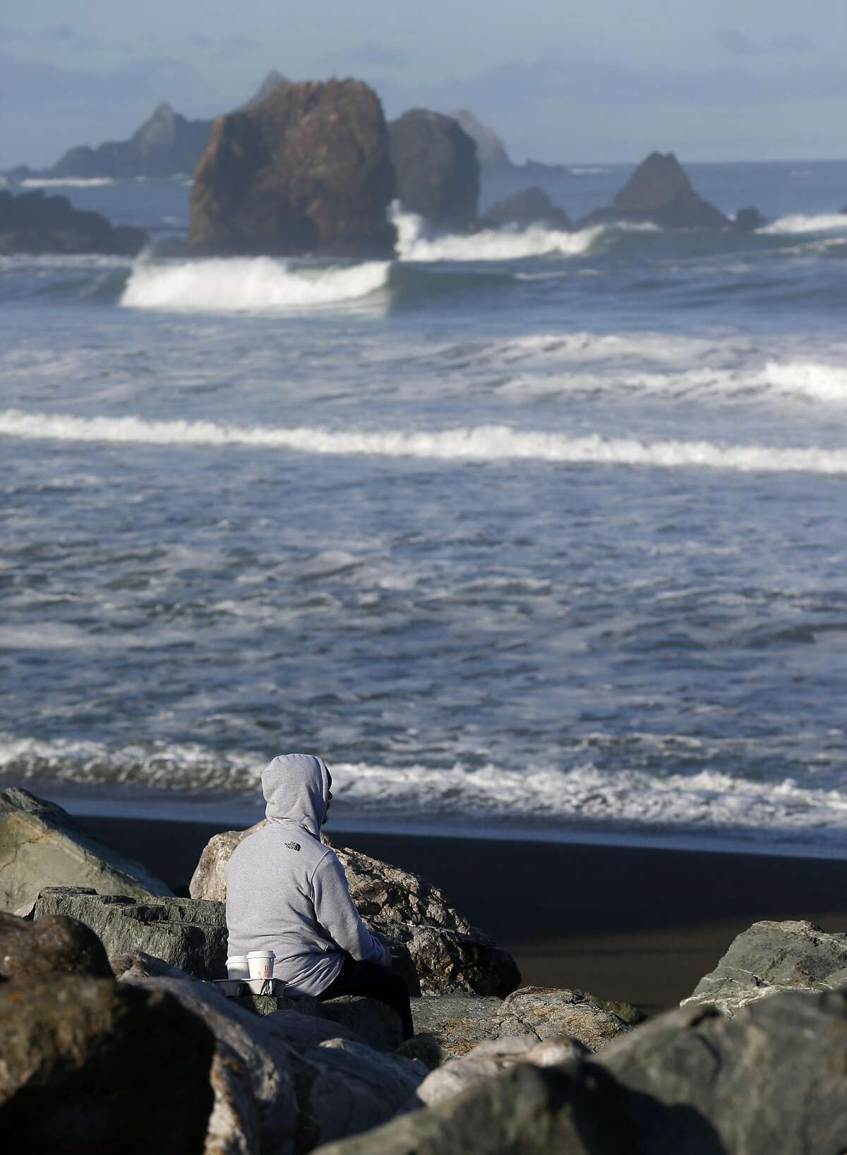 A man sits on the shore while the Coast Guard searches the ocean for a swimmer who was reported missing in the early morning hours off Rockaway Beach in Pacifica, Calif. on Tuesday, March 26, 2019.