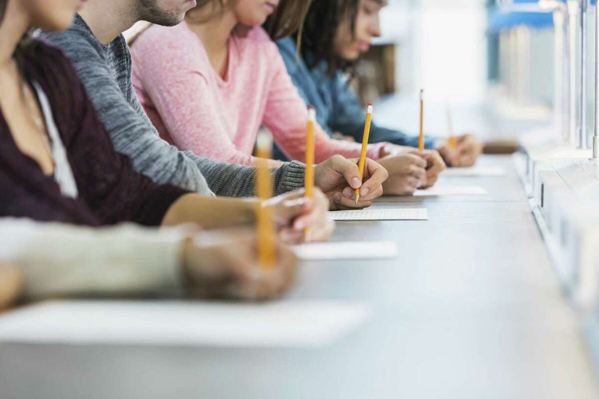 Cropped view of a multiracial group of young men and women sitting in a row at a table, writing with pencils on paper. They are taking a test or filling out an application. Focus is on the hand of the young man in the middle in the gray shirt.