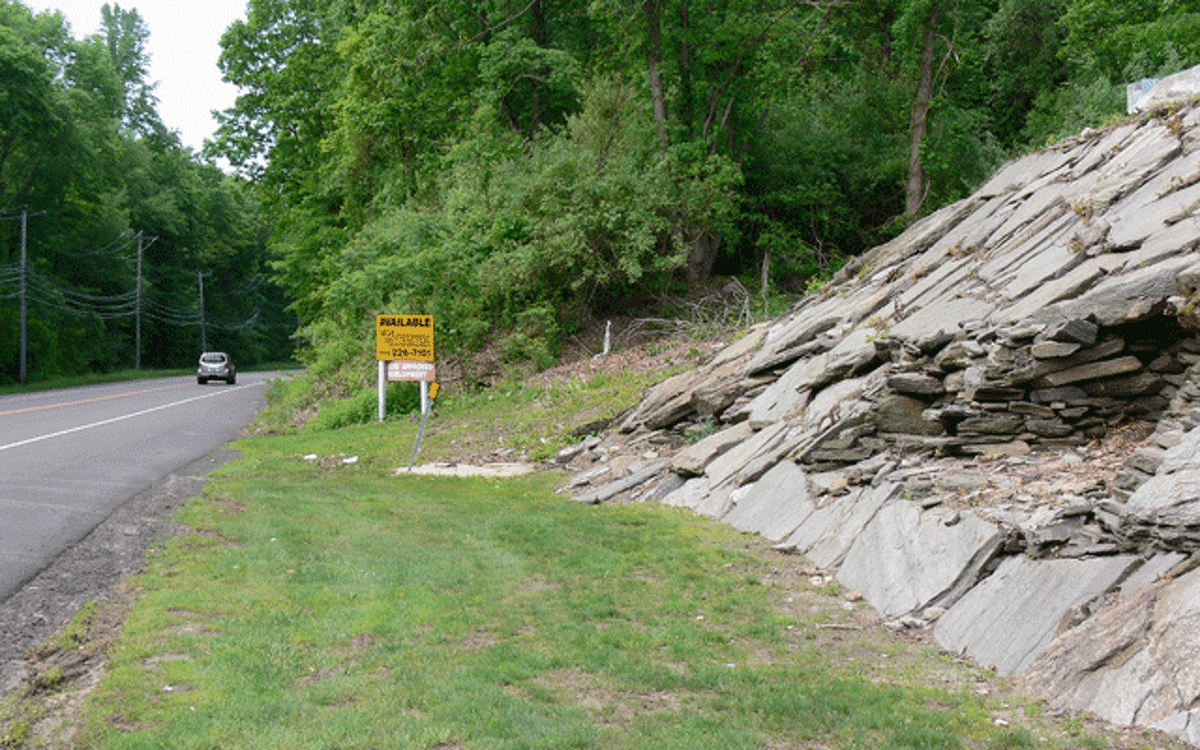The yellow “Available” sign indicates where the two-acre site begins, looking south on Bridgeport Avenue. The ledge area shown belongs to the adjoining lot, and the undeveloped parcel’s terrain is generally similar to that of the adjoining lot.