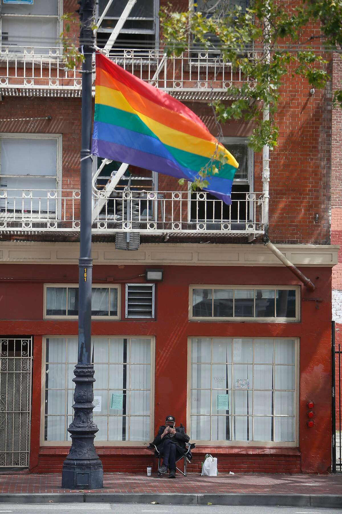 A man sits on Market Street below a rainbow flag and next to the Civic Center Navigation Center on Monday, June 24, 2019 in San Francisco, Calif.