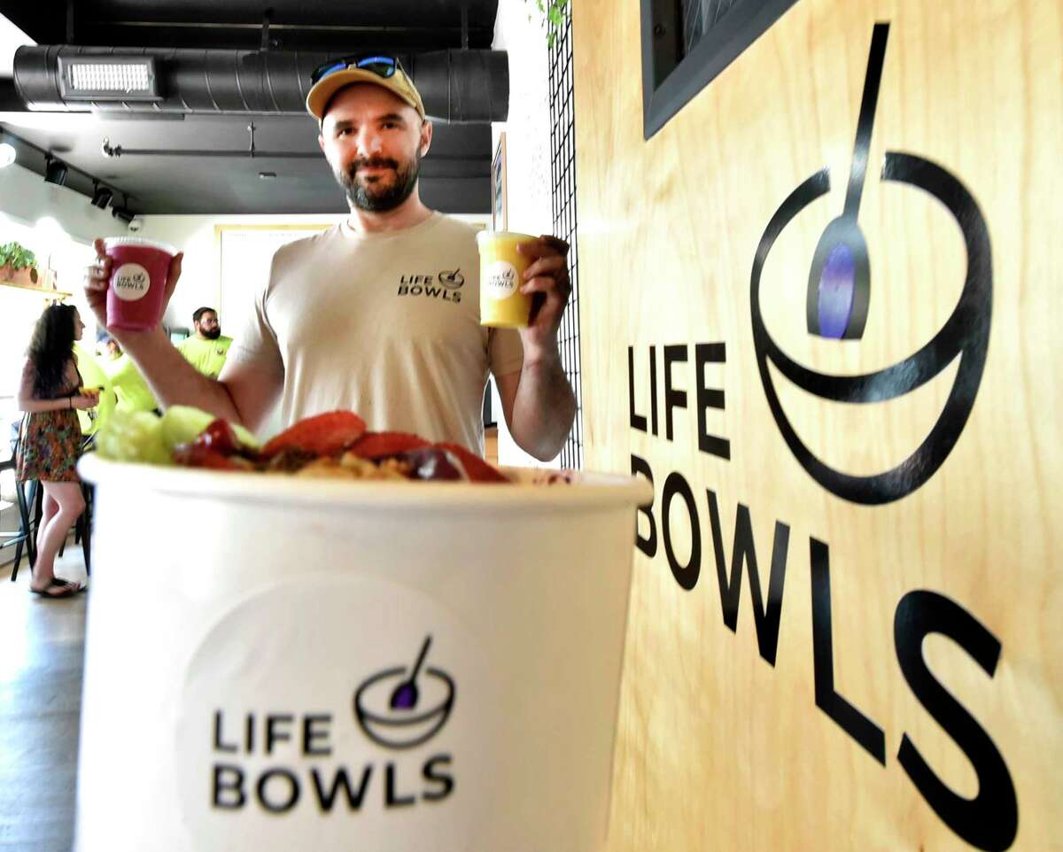 Madison, Connecticut - Friday, June 28, 2019: Co-owner Jonathan Bone, who with Justin Mclaughlin, created Life Bowls in Madison that offers Acai bowls, smoothies and fine coffee. Acai bowls have blended Acai berries from Brazil that is known to have a high content of antioxidants and no sugar. Jonathan holds a Pink Panther smoothie, left, and a Mango #5 smoothie, right. The Pink Panther is blended with pitaya, mango, strawberries, apple juice and almond milk with an option to include chia seeds. The Mango #5 smoothie ingredients are mango, pineapple, banana, honey, orange juice and as an option, coconut. The acai bowl, front, is "Kiwi's Playhouse" whose ingredients include a blend of acai, blueberries, mango pineapple, banana, lime, coconut water with toppings of kiwi, strawberries, chia seeds, granola, coconut, and honey.