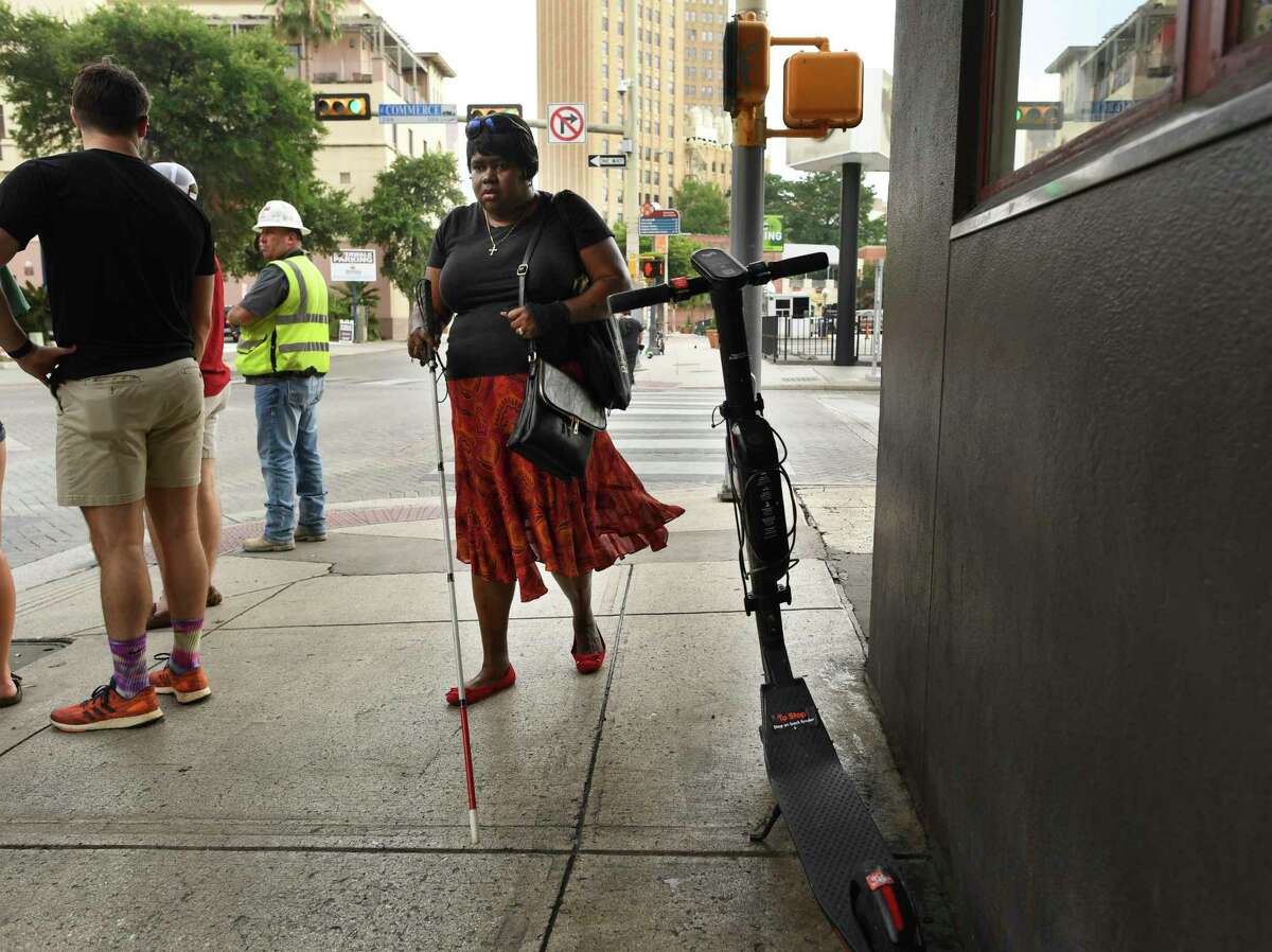 Athalie Malone, who is blind, walks by a scooter parked on a sidewalk along Navarro Street on her way to a city government meeting last month. Malone is on the selection committee that will determine which e-scooter companies will be awarded lucrative long-term city contracts, and her left arm is currently in a sling from her third downtown stumble this year over an abandoned scooter.