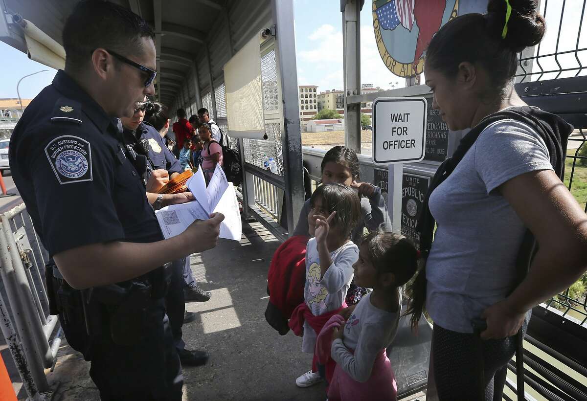 In this July 17, 2019 photo, a United States Customs and Border Protection Officer checks the documents of migrants, before being taken to apply for asylum in the United States, on the International Bridge 1 in Nuevo Laredo, Mexico. Mexico has received some 20,000 asylum seekers returned to await U.S. immigration court dates under the program colloquially known as "remain in Mexico." But there had been no sign of such large-scale moving of people away from the border before now, after the program expanded to Nuevo Laredo in violence- and cartel-plagued Tamaulipas, a state where the U.S. State Department warns against all travel due to kidnappings and other crime. (AP Photo/Marco Ugarte)