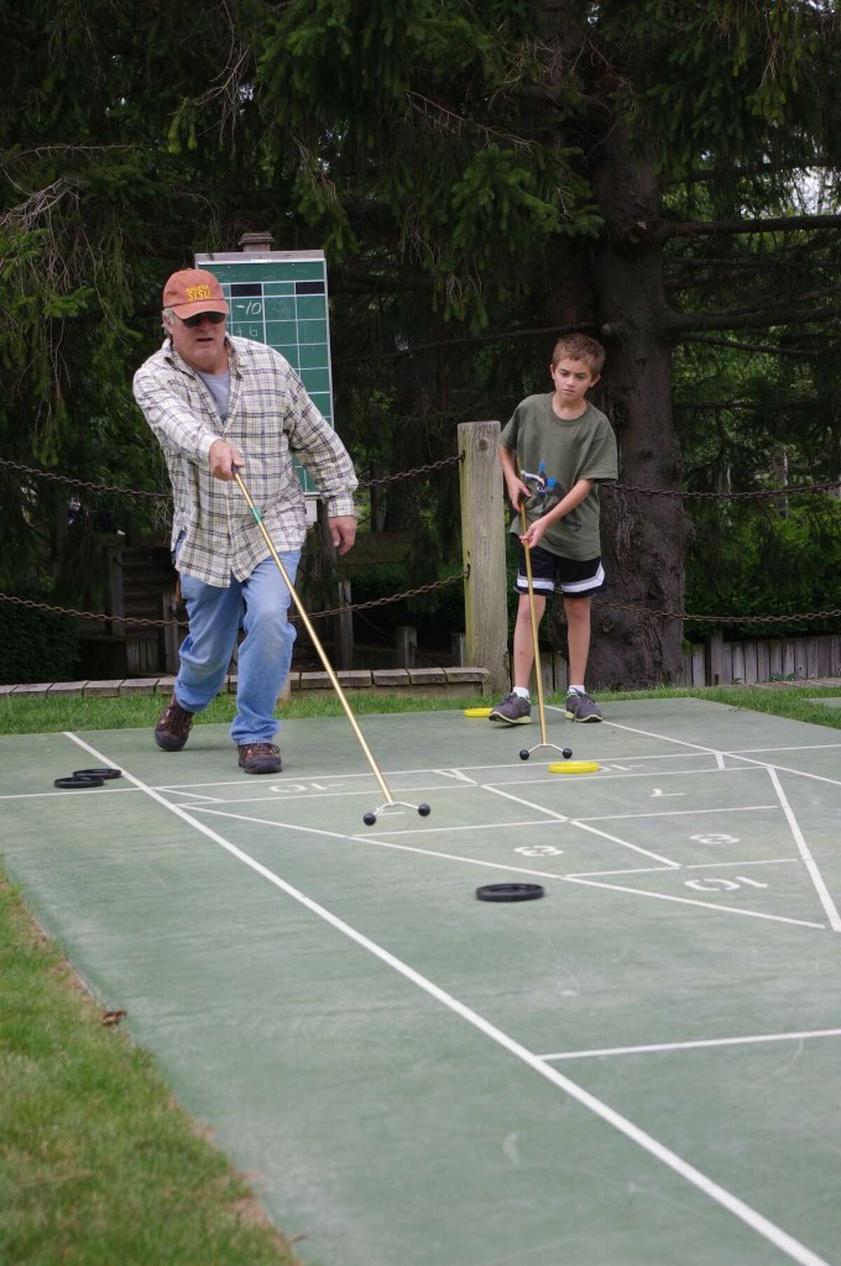 Gregg King uses a cue to shoot a shuffleboard disc toward the scoring diagram, while opponent Jack Huber watches. King would like to see more people use the courts that are located near the Manistee City Marina. (Dave Yarnell/News Advocate)