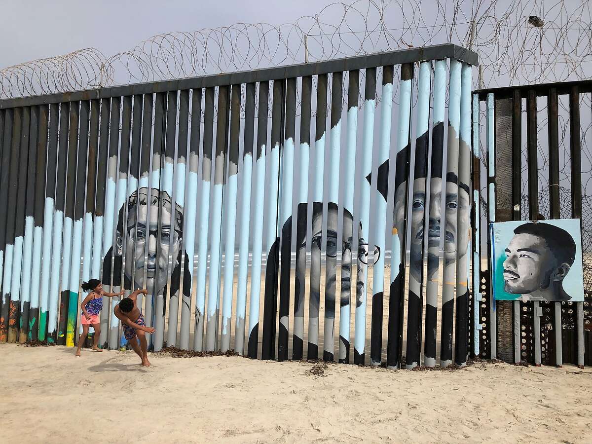 Children play in front of a new mural on the Mexican side of a border wall in Tijuana, Mexico Friday, Aug. 9, 2019. The mural shows faces of people deported from the U.S. with barcodes that activate first-person narratives on visitors' phones. Lizbeth De La Cruz Santana conceived the interactive mural in Tijuana as part of doctoral dissertation at the University of California, Davis. (AP Photo/Elliot Spagat)