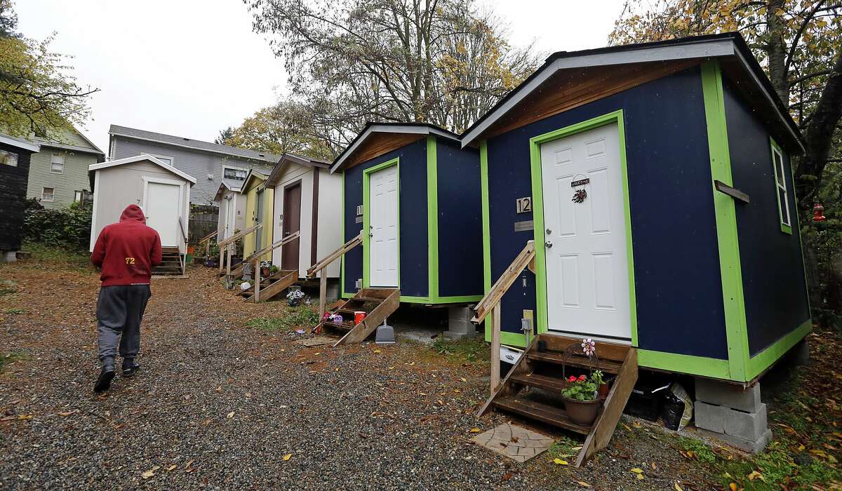 In this Nov. 9, 2017 file photo, a resident walks past a row of tiny houses at a homeless encampment in Seattle where full size homes stand behind. Seattle Mayor Jenny Durkan wants to move hundreds more homeless people into tiny homes, emergency shelters and other immediate housing in the next 90 days. (AP Photo/Elaine Thompson, file)