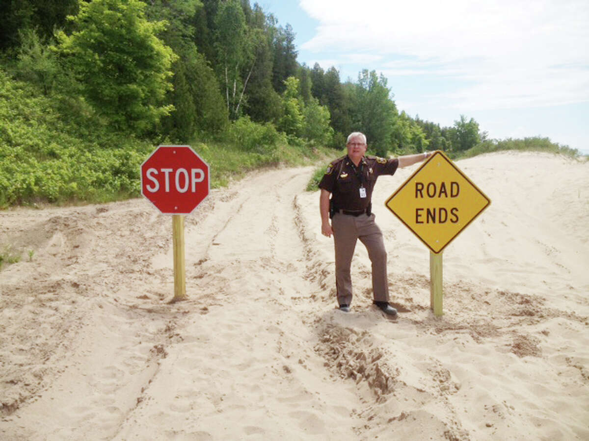 STOP HERE: Benzie County Sheriff Ted Schendel stands near signs posted at the end of Lakeside Boulevard, which mark where public access ends and private property begins. The sheriff’s office and the Department of Natural Resources will be actively ticketing people driving on the dunes, the beach or private property. (Courtesy Photo)