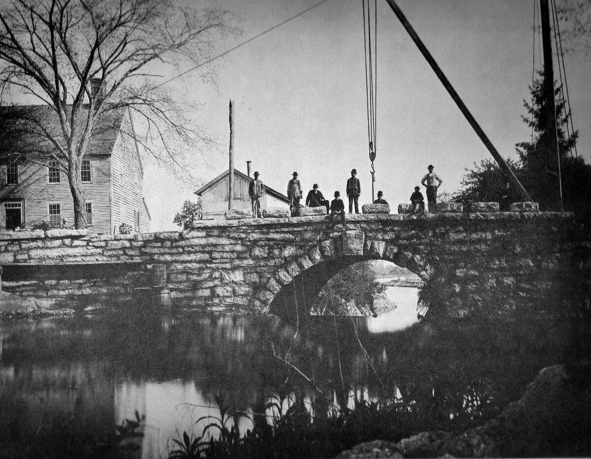 The building of Memorial Bridge, 1888-1889, by John Beatty of Leetes Island. The granite blocks that have the names of the first settlers are being set on the north coping with the aid of a hand-operated crane. The building to the left is the Fowler Homestead.