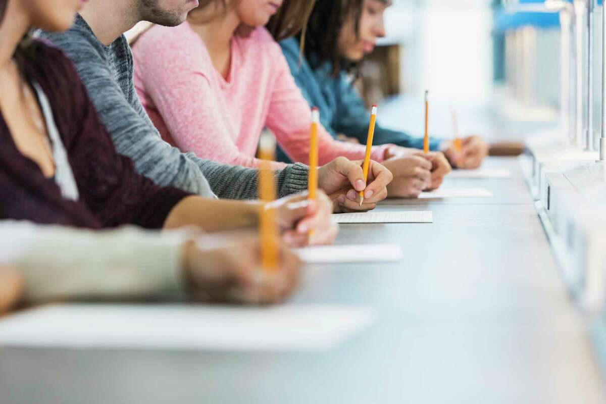 Cropped view of a multiracial group of young men and women sitting in a row at a table, writing with pencils on paper. They are taking a test or filling out an application. Focus is on the hand of the young man in the middle in the gray shirt.