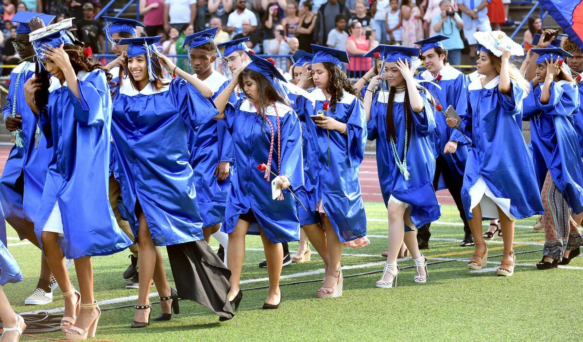 Graduates hold onto their caps as a gust of wind passes as they enter the football field for graduation at West Haven High School.