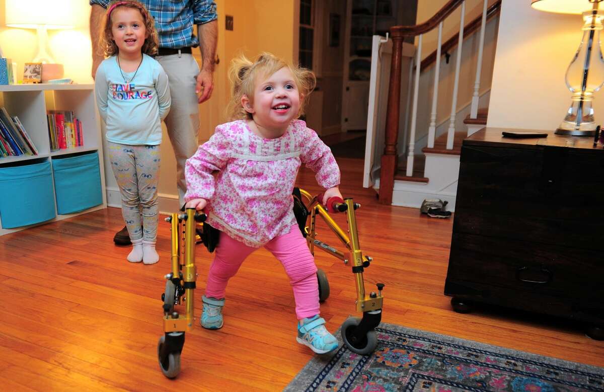 Serena Zitnay, 3, uses her walker to get around as her older sister Emma, 5, looks on while at home in Orange. The family has decided to raise money to try and have a walker made that is more kid friendly.