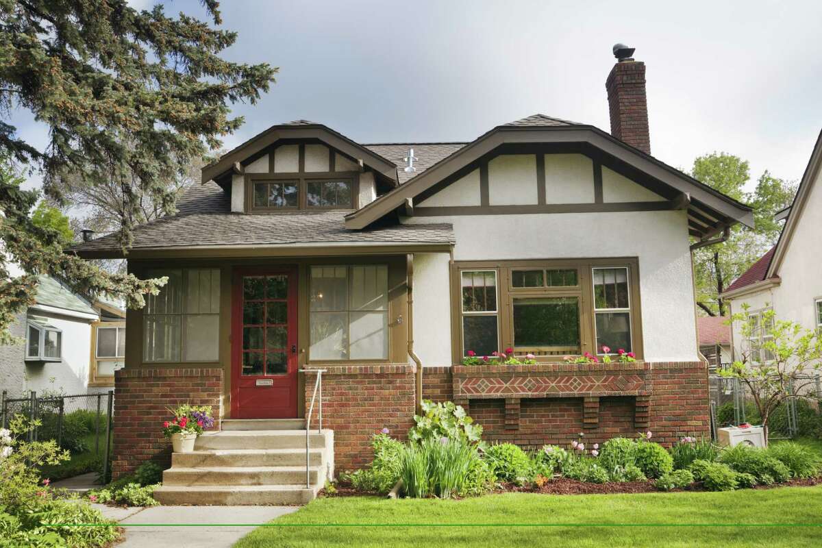 Full exterior front facade of 1920s era Arts and Crafts bungalow house. An example of Midwest, USA residential building style, the old, quaint architecture features stucco and brick walls, red front door, and picture window looking out to the lawn. A springtime flower garden grows outdoors. Horizontal, straight-on front view.