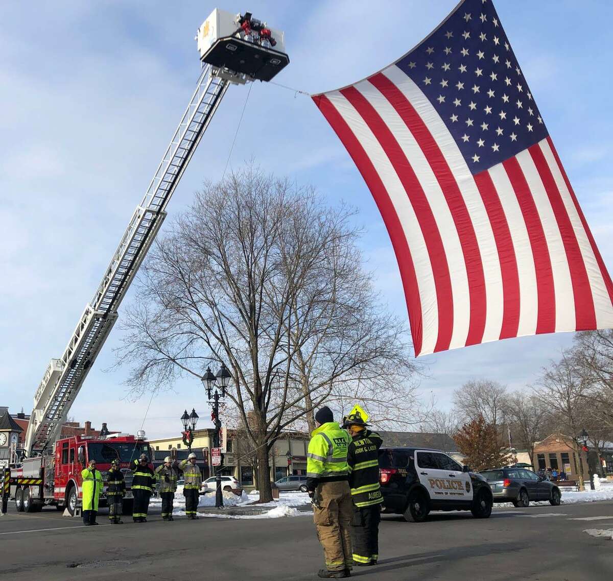 Spectrum/ A large American flag draped across the north side of Main Street in New Milford last Friday, Dec. 6, 2019, paid tribute to Ronald J. Adams of New Milford. Adams, 71, died Nov. 29 at his home. Firefighters from Water Witch Hose Co. #2 in New Milford and Newtown fire department hung the flag between New Milford’s Tower 25 and Newtown’s Truck 14 from the crossover near the bandstand to Church Street. A member of Iron Works Local #40, Adams had been of the construction of the World Trade Center. After 9/11, Adams stepped up and was part of the rescue/recovery efforts, which contributed to his disease. Above, Water Witch members, from left to right, on left, Bob Golembeski, Al Tani, David Golembeski, Lt. Sean Delaney and Chief Jim Ferlow, as vehicles in the procession make their way beneath the flag. New Milford Tower 25 and Newtown Truck 14