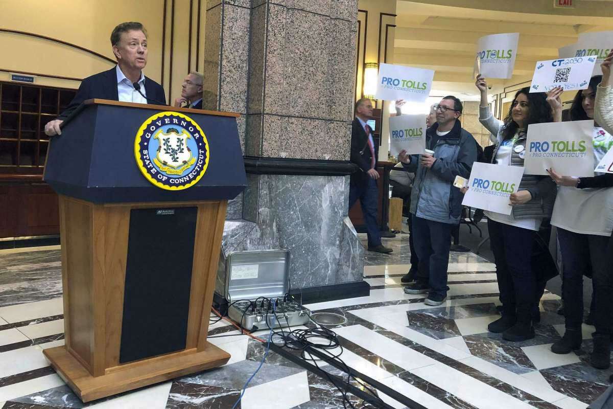 Gov. Ned Lamont answers questions from the media following a meeting of the State Bond Commission, Wednesday, Dec. 18, 2019, at the Legislative Office Building in Hartford, Conn. A group of mostly Fairfield County residents hold signs showing their support for tolls.