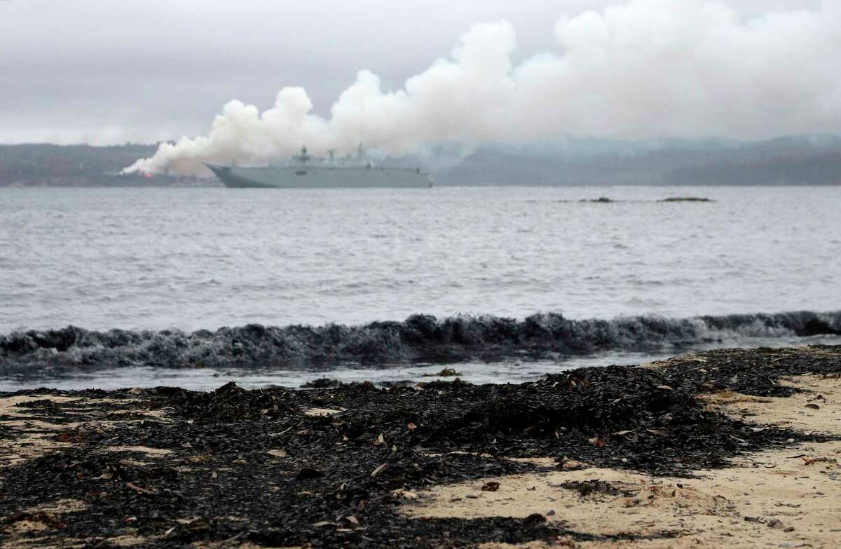 A beach is littered with charred leaves as a huge pile of wood chips burn at a mill across the bay with HMAS Adelaide standing by to assist at Eden, Australia, Thursday, Jan. 9, 2020. Wildfires have destroyed 2,000 homes and continue to burn, threatening to flare up again as temperatures rise. (AP Photo/Rick Rycroft)