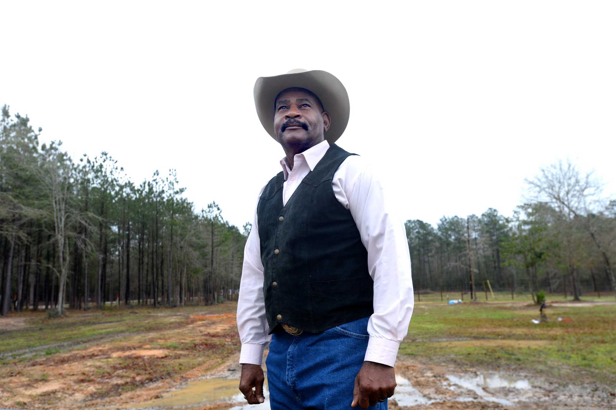 Guy is standing and looking straight on water and trees. He wears vest, cap  and shirt.