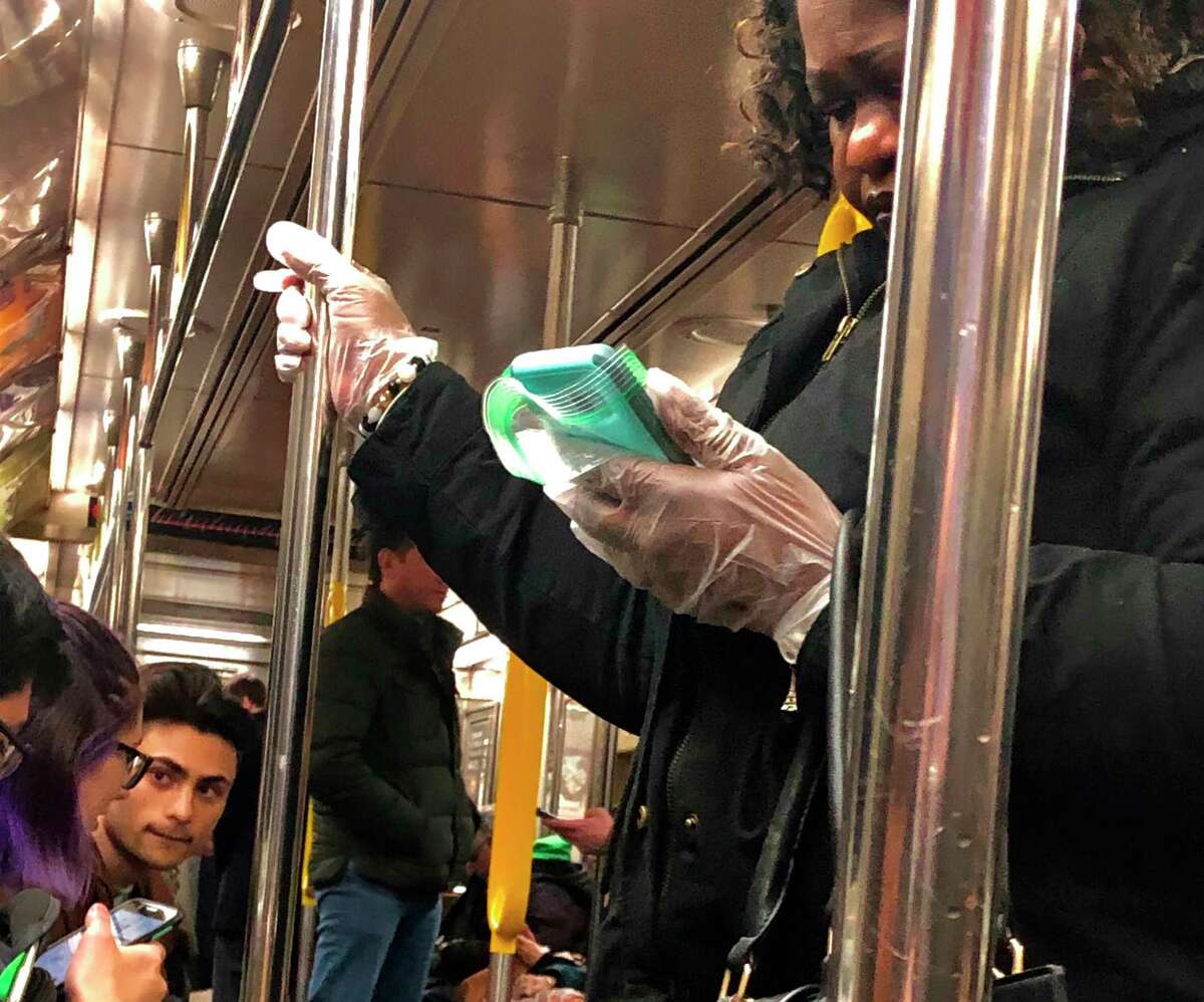 A woman uses protective gloves as she looks at her phone wrapped in a plastic bag while riding a New York City subway train, Monday, March 9, 2020.