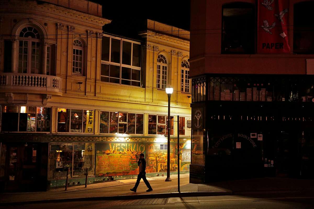 A pedestrian walks by darkened North Beach mainstays Cafe Vesuvio and City Lights Bookstore as the city began to shut down following an order to shelter in place in San Francisco, Calif., on Monday, March 16, 2020. The six Bay Area counties issued a shelter in place order for residents to try and curtail the spread of the Covid-19 virus.