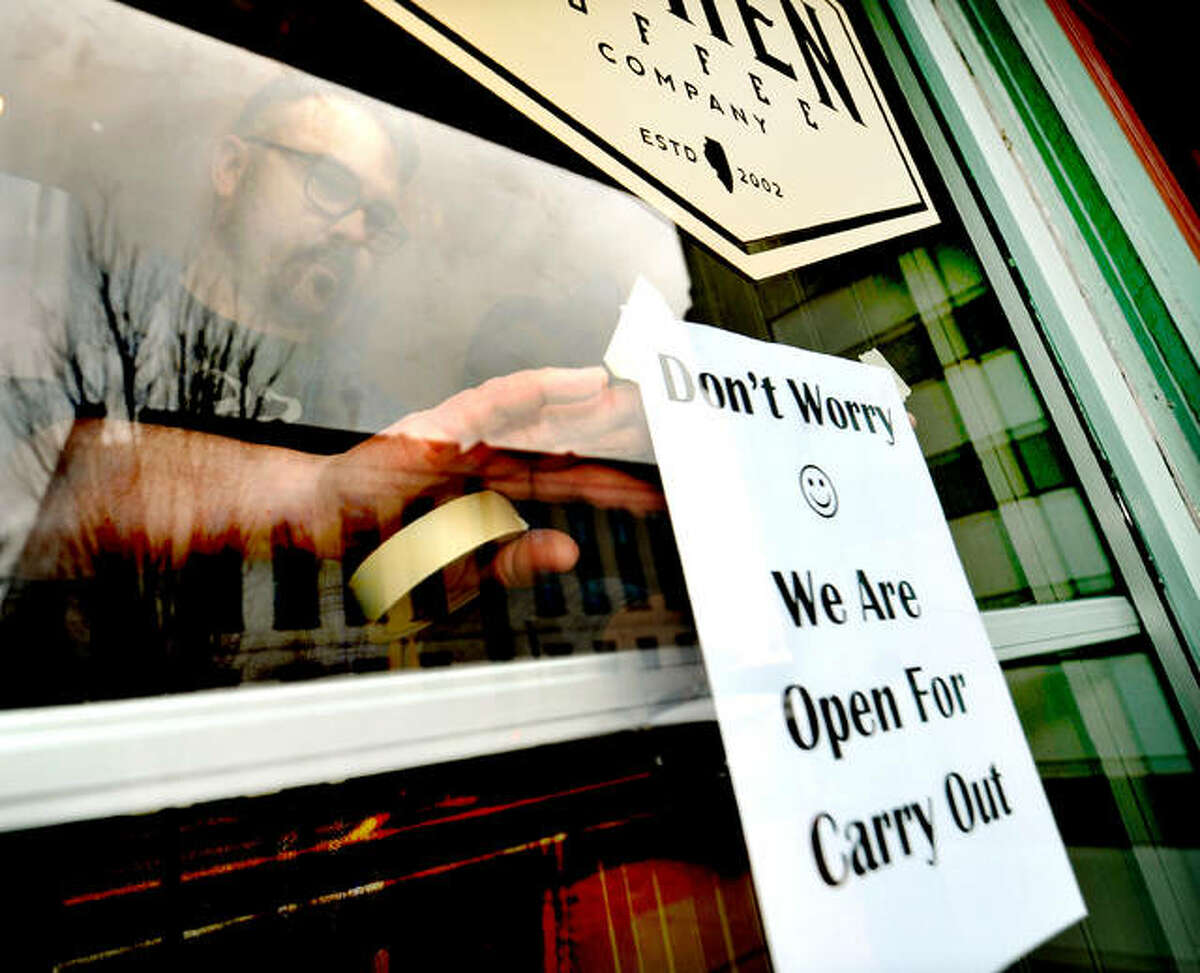 Trevor Taynor of 222 Artisan Bakery and Cafe hangs a sign on the front door Monday letting customers know that even though the dining room will be closed due to the coronavirus the store will still be open for takeout.