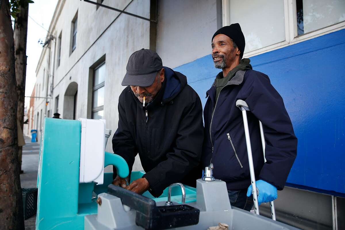 Brian Hayes (right) uses a foot pedal to pump water from a tap to help Hanna Wallace (left) wash his hands at a hand washing station outside of Multi-Service Center South on Thursday, March 19, 2020 in San Francisco, Calif.