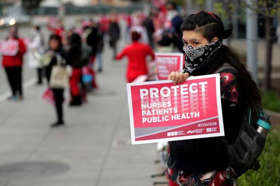 Valeria Vital, an ER tech, stands holding a sign during a protest by medical professionals working for Kaiser Permanente in Oakland and their supporters in Oakland, Calif., on Monday, March 23, 2020. The medical care professionals hoped to emphasize their need for better personal protective equipment (PPE) rather than having to use bandanas instead as they say Kaiser has asked them to do.