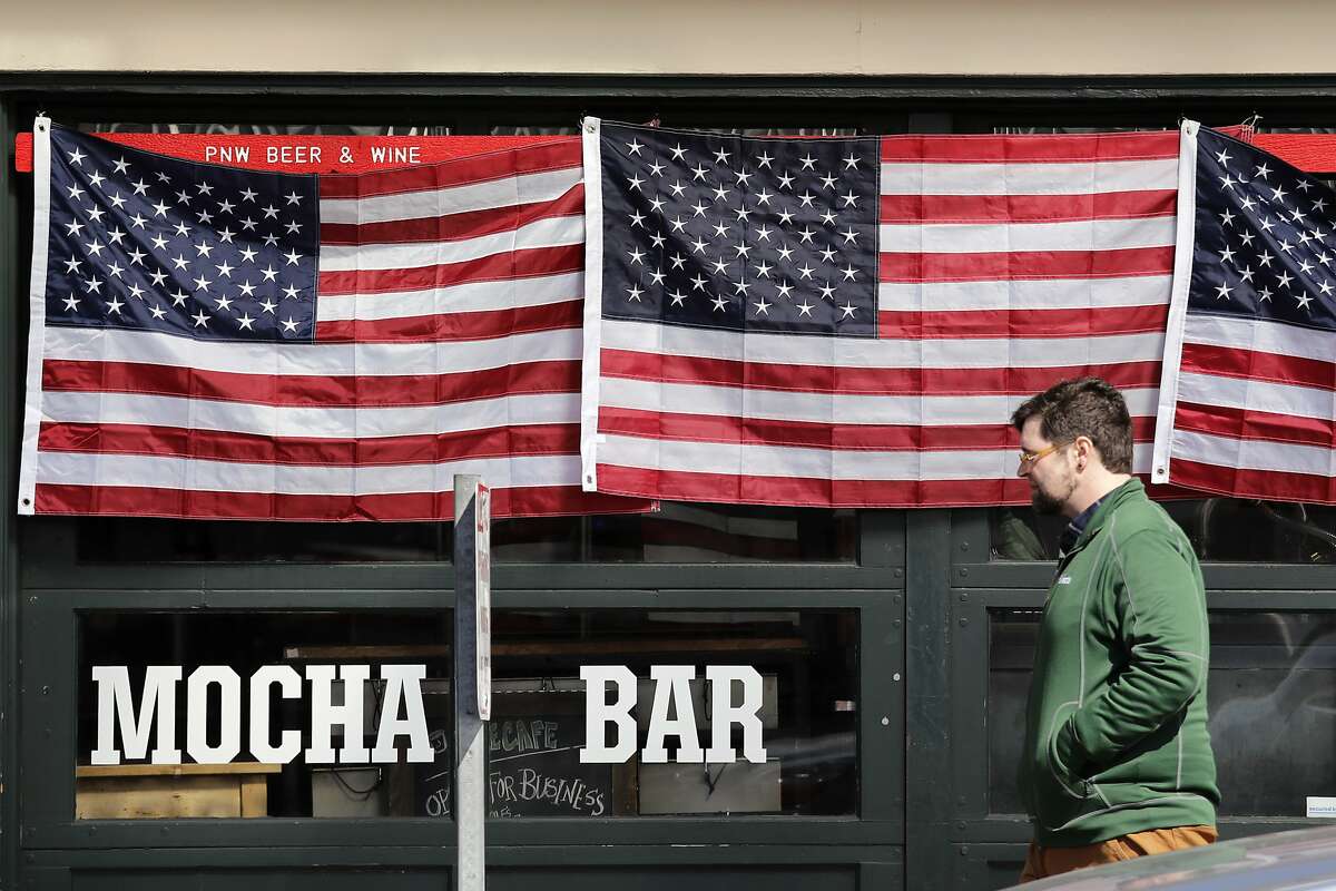 A pedestrian walks past a closed cafe with its windows covered in American flags at the Pike Place Market Friday, March 20, 2020, in Seattle. Restaurants, except for take-out orders, are closed, workers who can are working from home and people are being asked to maintain physical distance from others to help stop the spread of COVID-19. Washington state health officials reported eight new coronavirus deaths on Friday, bringing the total to 83. Seven of those deaths were in King County, the epicenter of the outbreak in the state. (AP Photo/Elaine Thompson)