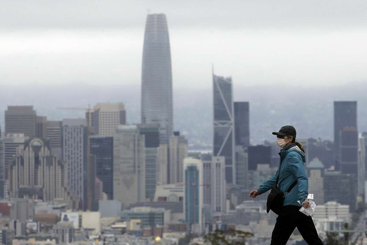 A woman wears a mask during the coronavirus outbreak while crossing a street in front of the skyline in San Francisco, Saturday, April 4, 2020.