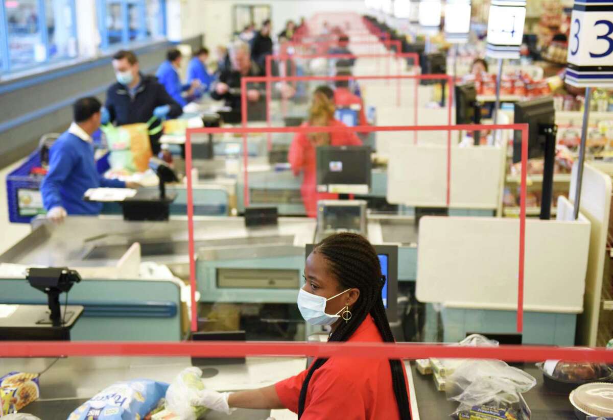 A sheet of plexiglass separates cashier Nadine Colas from customers in line at ShopRite of Commerce St. in Stamford, Conn. Monday, March 30, 2020. Many grocery stores are taking precautions in preventing the spread of coronavirus including signs promoting social distancing and plexiglass between shoppers and cashiers.