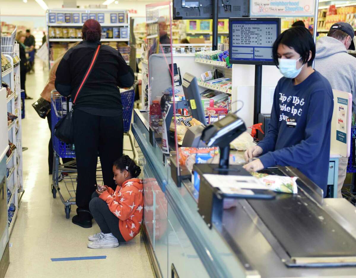 Greenwich's Ezell Pirtle, 6, plays a game while waiting behind a line of blue tape to check out with her motheras a sheet of plexiglass separates cashier Miguel Hernandez from customers in line at ShopRite of Commerce St. in Stamford, Conn. Monday, March 30, 2020. Many grocery stores are taking precautions in preventing the spread of coronavirus including signs promoting social distancing and plexiglass between shoppers and cashiers.