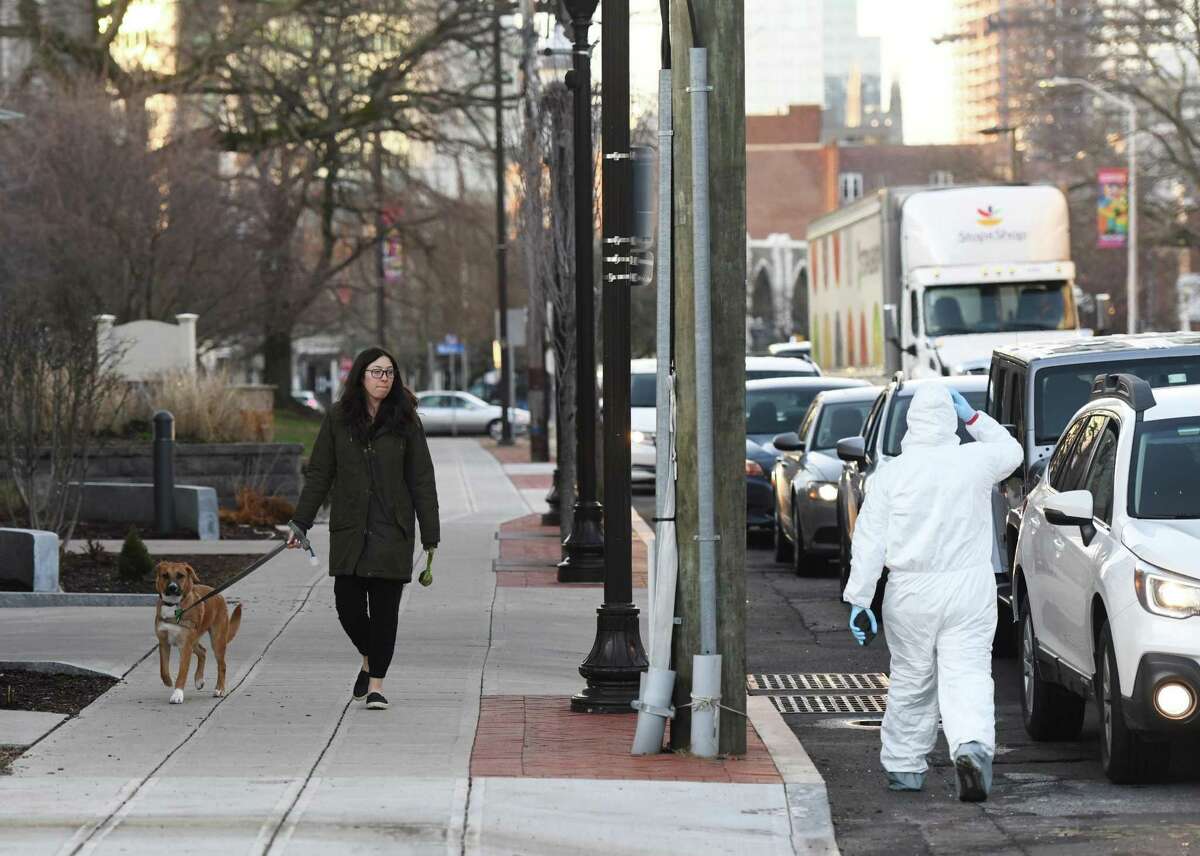 Katie Reitman walks her dog, Kona, past the line of cars waiting to be screened for coronavirus at the Murphy Medical Associates testing site on Bedford Street in Stamford, Conn. Wednesday, March 18, 2020. Murphy Medical Associates has been offering drive-thru testing for the coronavirus at locations in Greenwich, Stamford, Darien, New Canaan, and Stratford. An appointment must be approved and booked in advance to be tested.