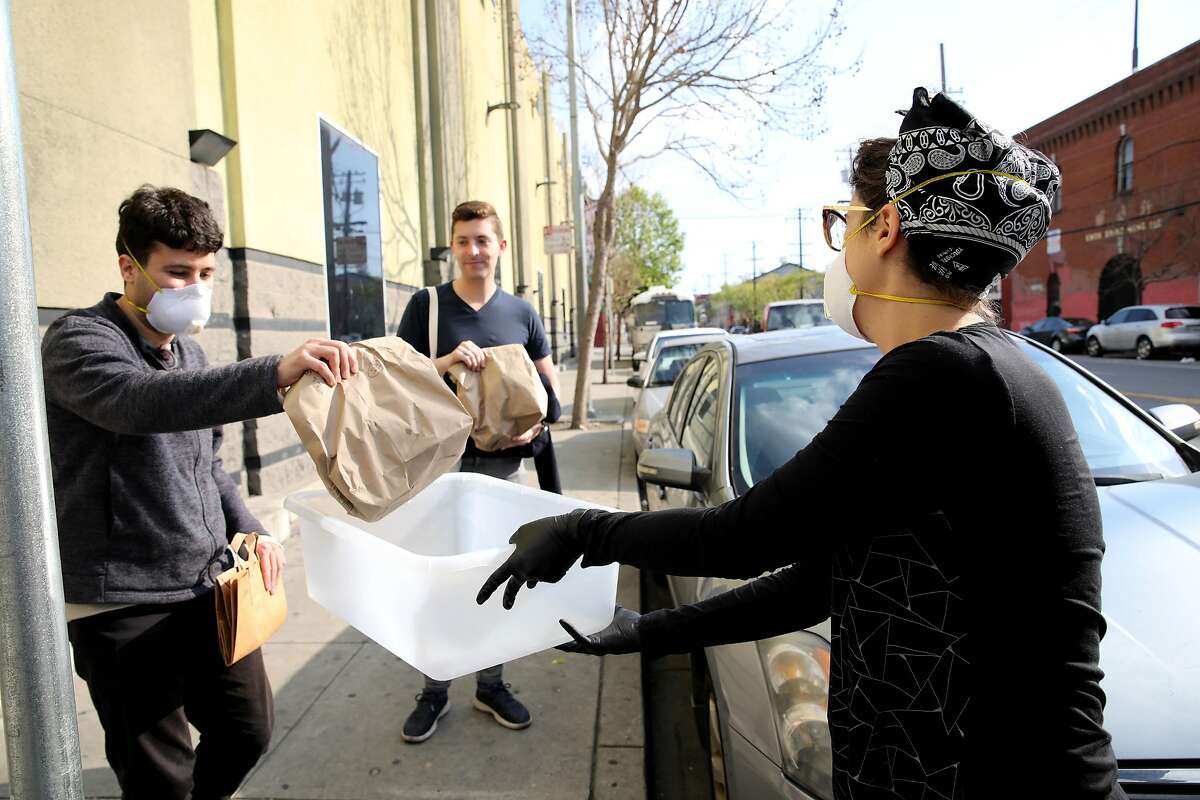 Ben Hiller, left, and Jacob Hurwitz, center, pick up their food orders from Rachel Metcalf, right, an employee with SM Vegan, outside of the closed Best Buy at Harrison and 14th St. on Friday, March 20, 2020, in San Francisco, Calif. "We're trying to make people feel comfortable as long as possible," Metcalf said.