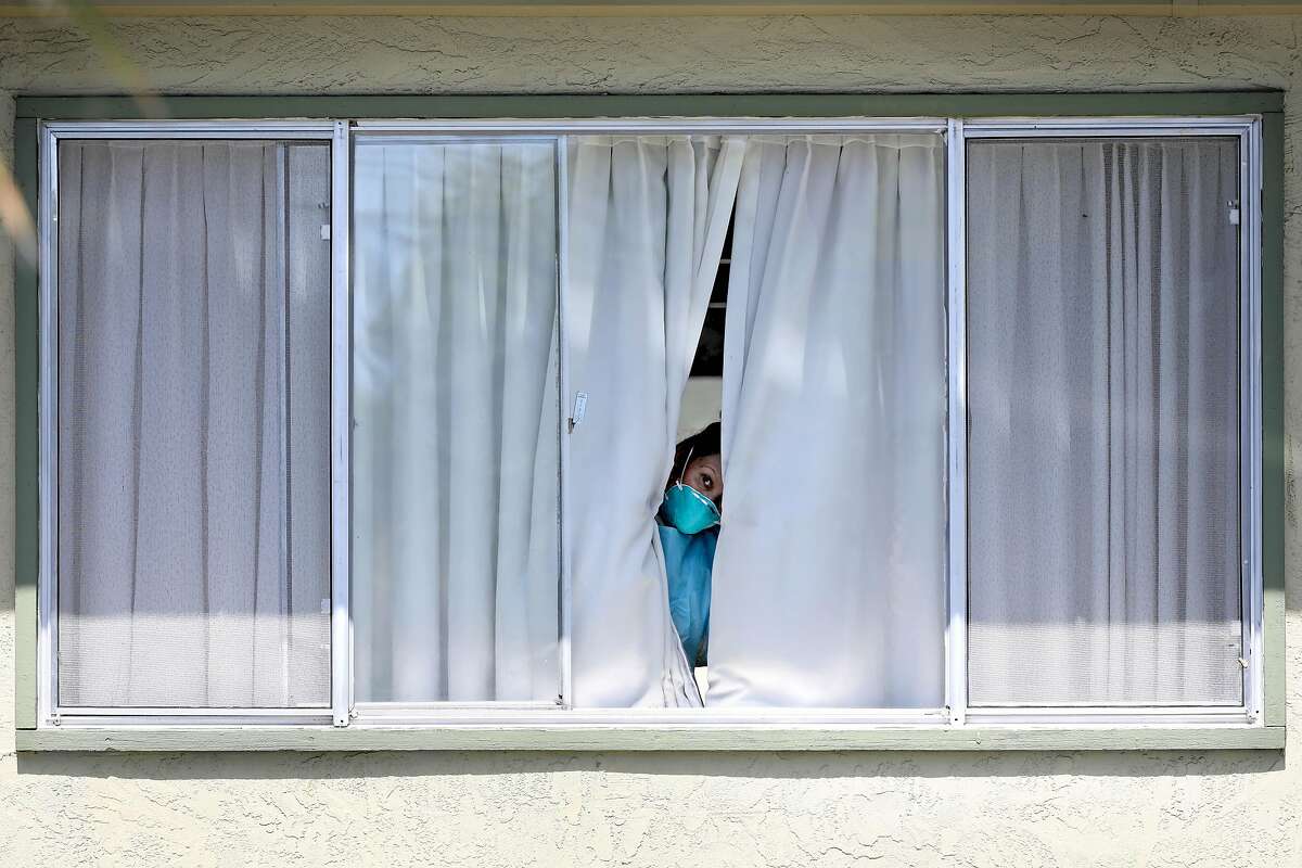 An employee wearing protective gear peeks from a window after a patient was picked up by Falcon Critical Care Transport at Gateway Care and Rehabilitation on Wednesday, April 15, 2020, in Hayward, Calif. The facility currently has eleven COVID-19 related deaths with dozens of staff members and patients infected with the novel coronavirus.