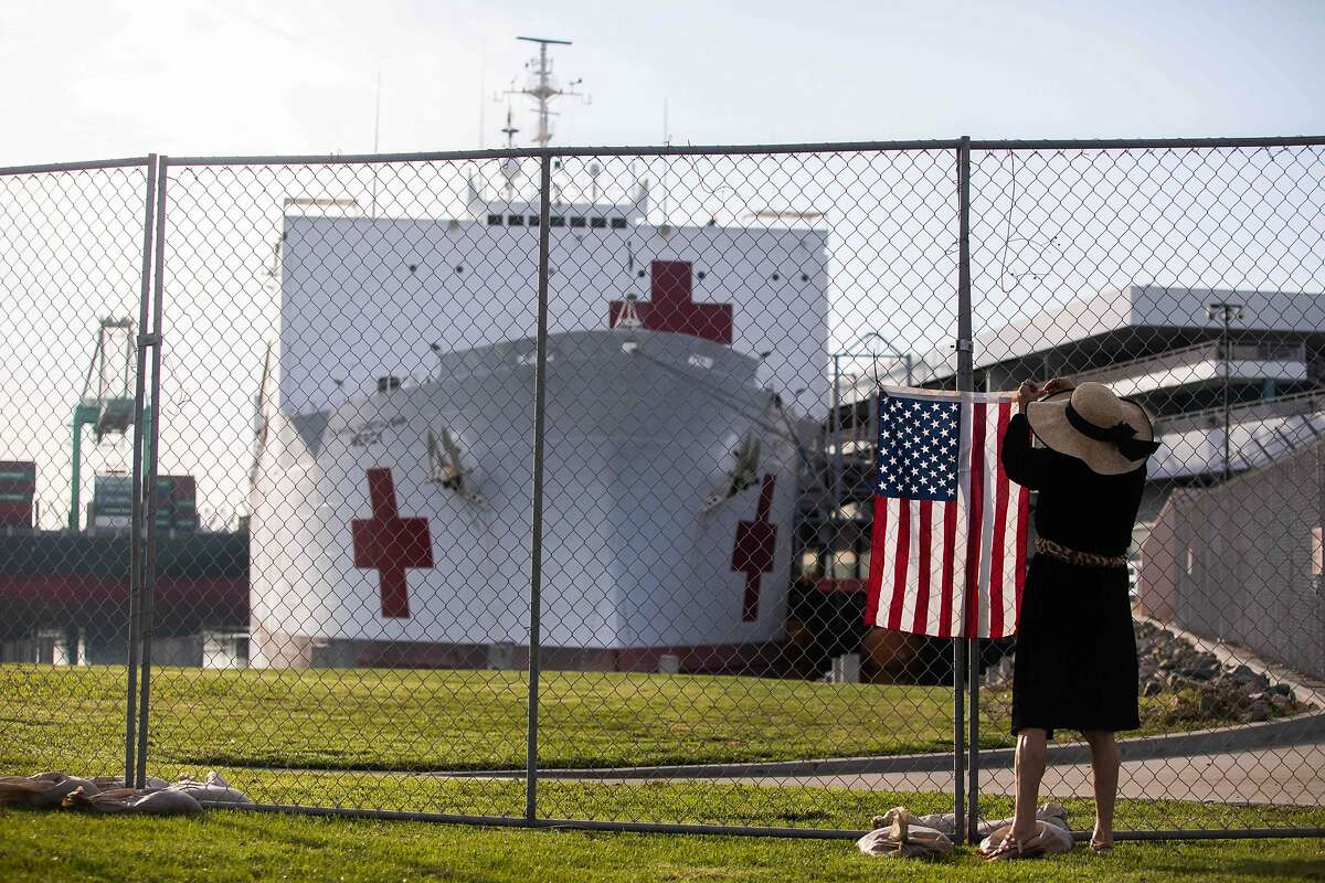 A woman hangs a US flag in a fence in front of the US Navy Hospital ship Mercy on March 28, 2020 at the Port of Los Angeles in the city of San Pedro to help local hospitals amid the growing Coronavirus crises in Los Angeles, California. - The ship hold 1000 beds which will be used to treat non-coronavirus patients. (Photo by Apu GOMES / AFP) (Photo by APU GOMES/AFP via Getty Images)