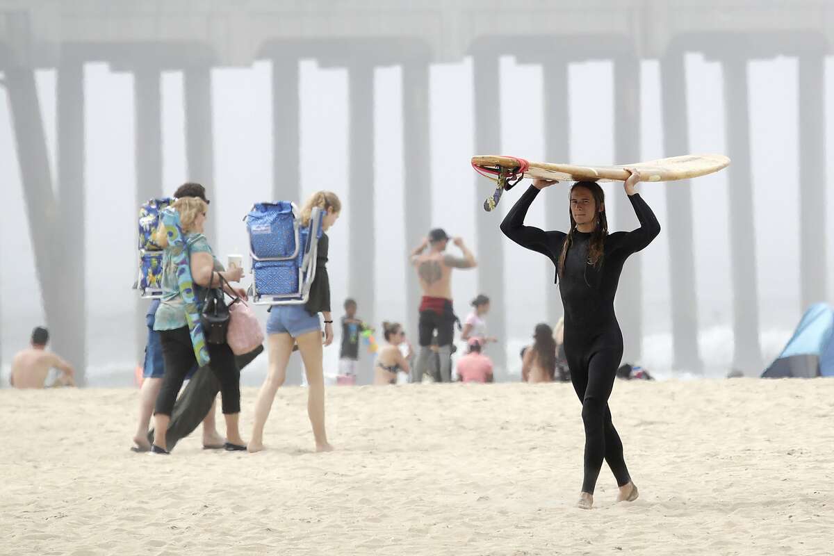 A surfer carries a board Sunday, April 26, 2020, in Huntington Beach, Calif. A lingering heat wave lured people to California beaches, rivers and trails again Sunday, prompting warnings from officials that defiance of stay-at-home orders could reverse progress and bring the coronavirus surging back. (AP Photo/Marcio Jose Sanchez)