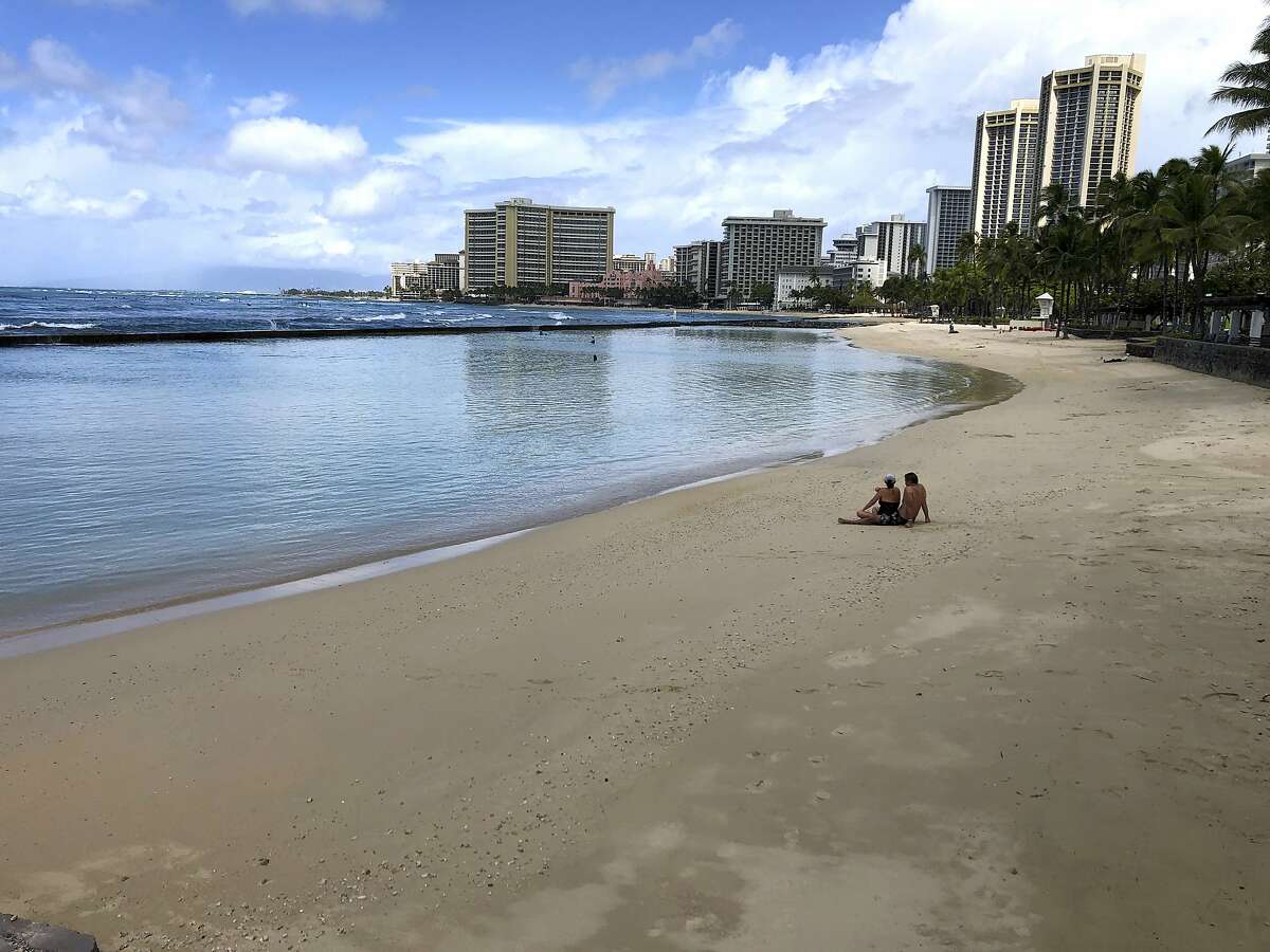 A couple sits on an empty section of Waikiki Beach in Honolulu. Hawaii law enforcement authorities are cracking down on rogue tourists who are visiting beaches, jetskiing, shopping and generally flouting strict requirements that they quarantine for 14 days after arriving. (AP Photo/Caleb Jones, File)