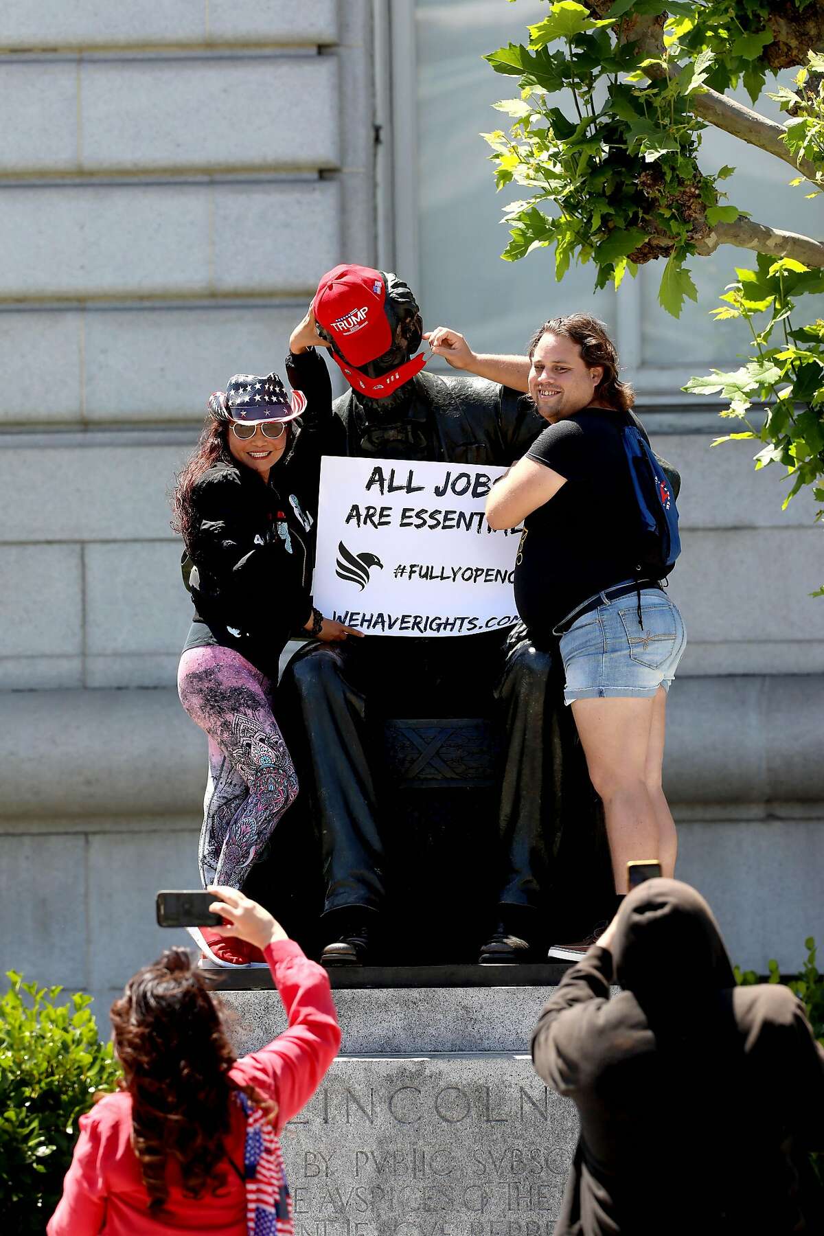Louisa Ip, left, and Jacob Sickness, pose for photographs while placing a Trump 2020 cap on the statue depicting former United States president Abraham Lincoln as they protest during a rally in front of City Hall calling for Gov. Newsom to immediately reopen California completely in San Francisco, Calif., on Saturday, May 9, 2020. Protesters argued they've been stripped of their rights. "The whole thing is a plandemic," Ip said. "My belief is the whole thing is planned to take our privacy away, plus they wanted to take our constitutional rights away." After climbing down the statue Sockness said, "I'm trying to get my freedom back."