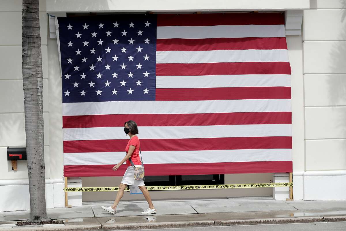 A woman wearing a protective face mask walks past an American flag in front of a closed business during the new coronavirus pandemic, Monday, May 11, 2020, in Palm Beach, Fla. Palm Beach County was authorized by Florida Gov. Ron DeSantis to initiate Phase 1 reopening regulations Monday, which includes limited reopening of retail establishments. (AP Photo/Lynne Sladky)