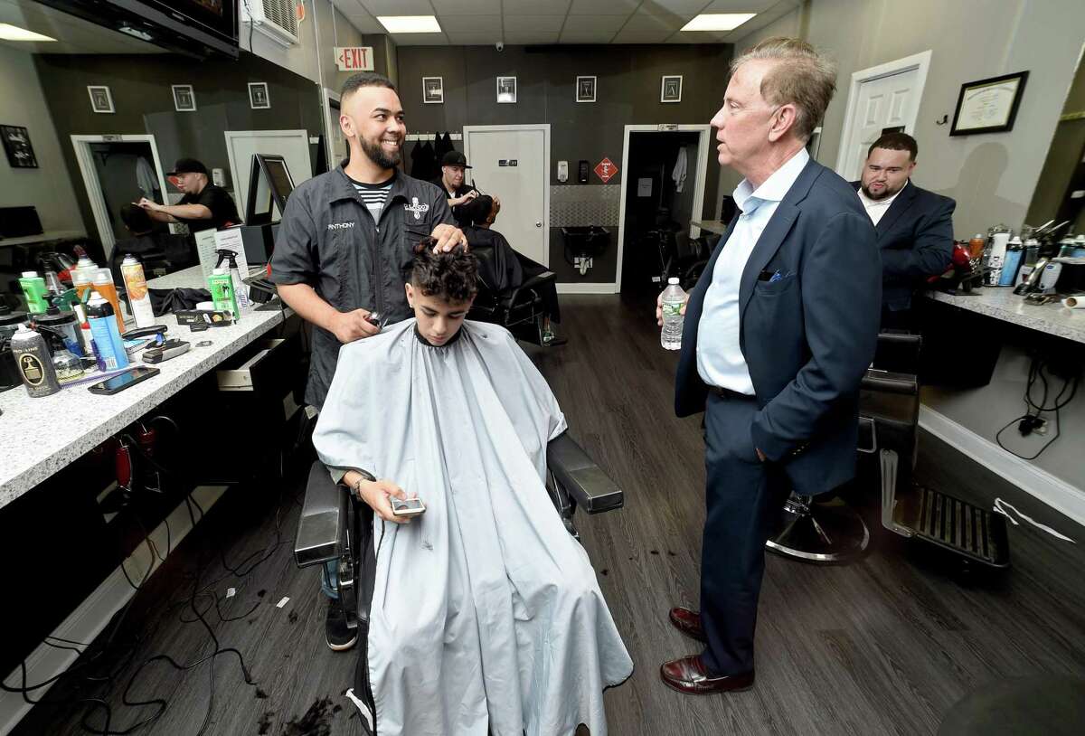 Then-candidate, now-Gov. Ned Lamont talks with Anthony Rivera (left) while Rivera cuts the hair of Alejandro Sanchez of East Haven at Orlando's Barber Shop on Grand Avenue in Fair Haven in 2018. On Monday, Lamont delayed the reopening of hair salons and barbershops to June 1, just two days before the scheduled restart.