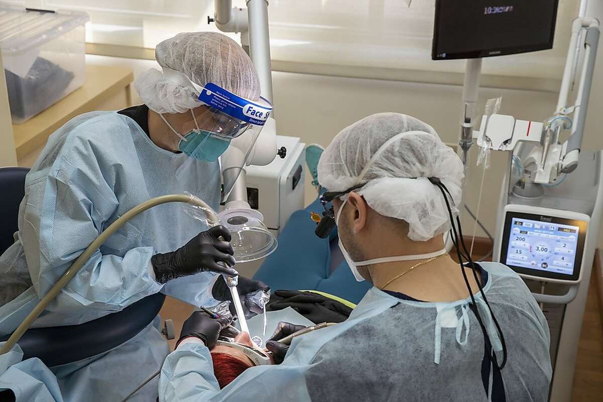 From left: Liv Tammik and Lior Tamir progress through a dental procedure on a patient at Bloom Dental Group on Friday, May 15, 2020, in San Mateo, Calif. The dental office has upgraded its safety, amid the coronavirus pandemic. It has several high-efficiency particulate air (HEPA) purifier and a device near the patient that continuously vacuums the surrounding air. The dentists have also upgraded their masks. They now wear N95 masks and face shields during procedures.