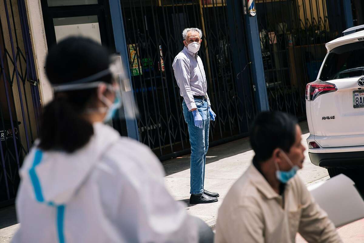 A man watches from a distance as health workers prepare nasal swab tests for the COVID-19 coronavirus disease for residents of the Hoy Sun Ning Yung Benevolent Association on Chinatown's Waverly Place in San Francisco, Calif. on Friday, May 22, 2020.
