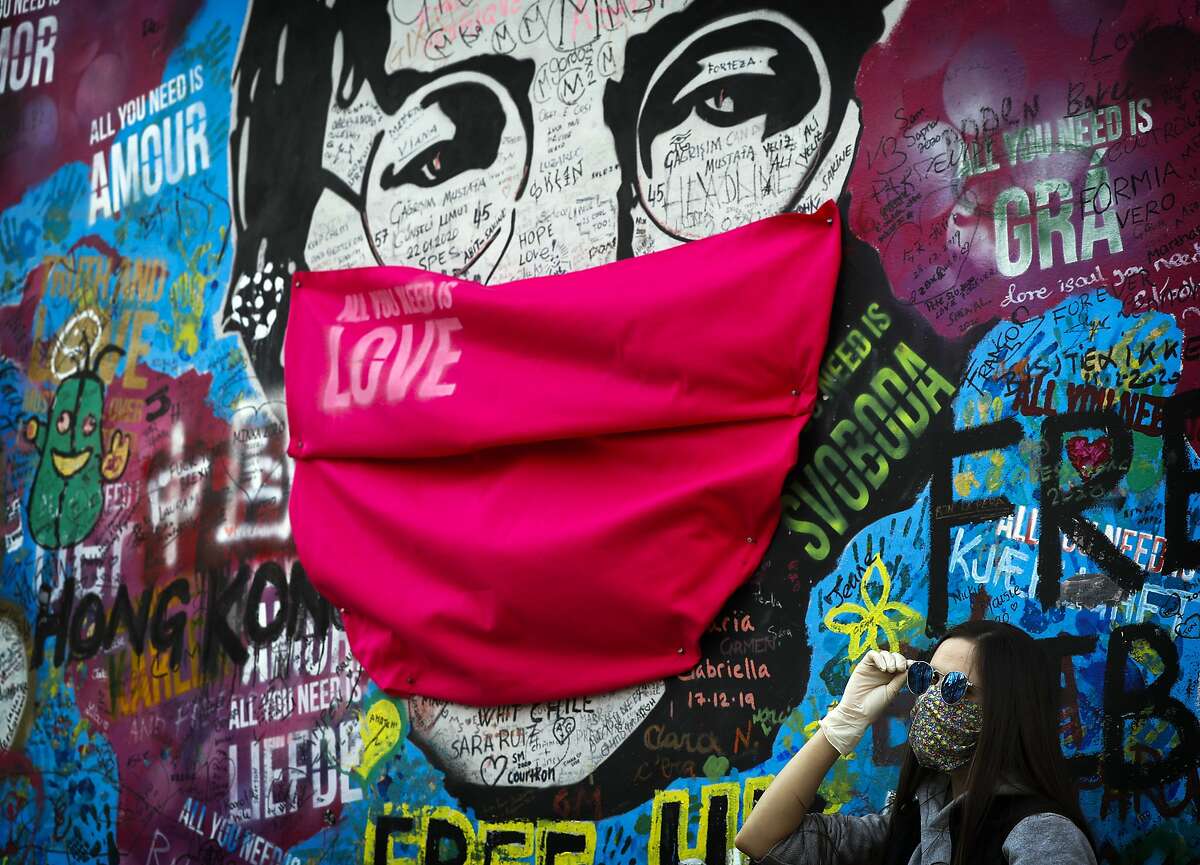 A woman poses for a photo by the "Lennon Wall" with a face mask attached to the image of John Lennon. (AP Photo/Petr David Josek)