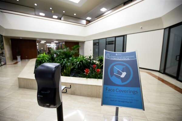 A sign requiring face coverings stands next to a hand sanitizer dispenser is in the lobby of the building where Foster Marketing has its office on Wednesday, May 27, 2020 in Houston. Foster Marketing is one of the offices that felt comfortable returning to the office, in part because the office is so spacious and they have their own offices.