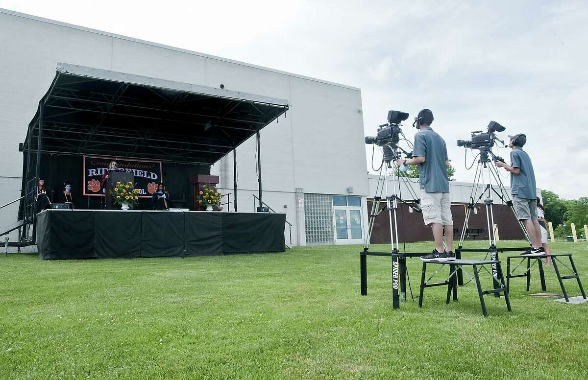 The Ridgefield High School graduation ceremony being broadcast to big screens in the school parking lot for graduates in their cars. Thursday, June 18, 2020. RIDGEFIELD - Seniors in black caps and gowns with Tiger-orange trim wandered a full school parking lot as they and their families prepared to watch graduation ceremonies on large screens. Ridgefield High School's 380 seniors graduated in two half-class sessions Thursday afternoon - commencement in the age of coronavirus.