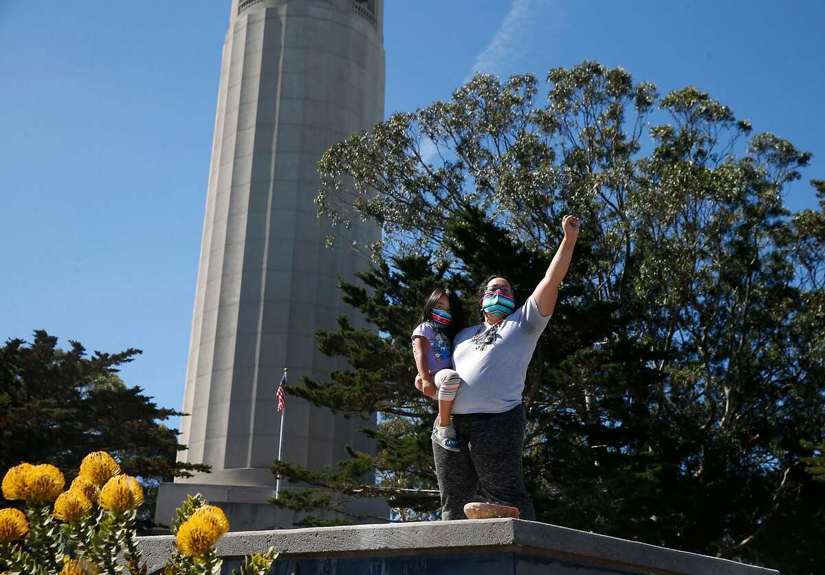 Mariposa Villaluna stands on a pedestal with their 4-year-old child in the parking lot at Coit Tower in San Francisco, Calif. on Thursday, June 18, 2020 after a crew from the city dismantled a statue of Christopher Columbus during the night.