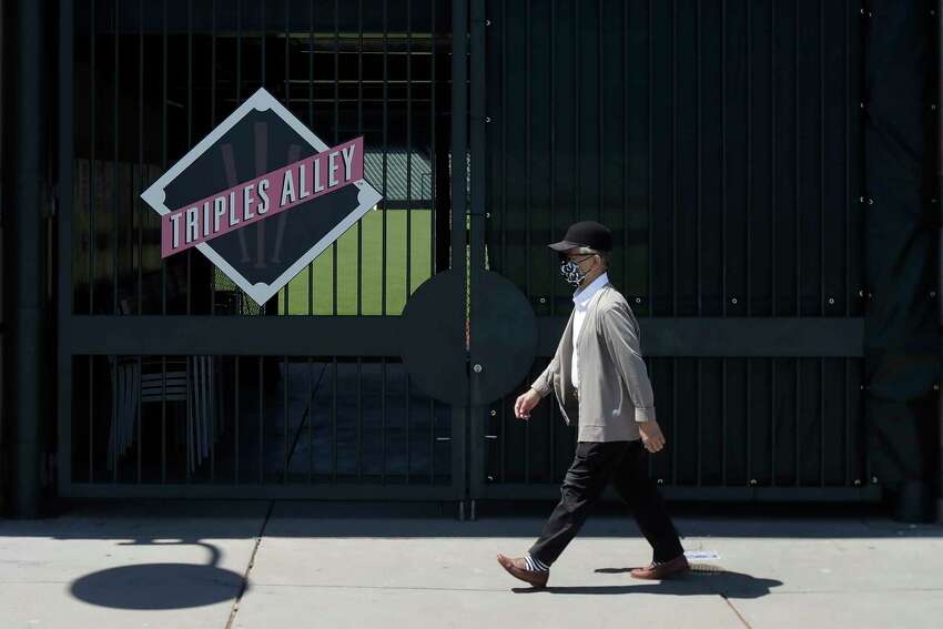 In his photo taken June 25, 2020, man wears a face mask during the coronavirus outbreak while walking past partially covered fencing outside Oracle Park, the San Francisco Giants' baseball ballpark, in San Francisco. This week, Major League Baseball players and owners reached an agreement to play an abbreviated, 60-game season that would start July 23 or 24 in teamsa€™ home ballparks. But the seats will be empty. Instead, fans hoping to see a game in person will be have to settle for pressing their faces up against hotel windows, squinting through metal grates or climb to rooftops when baseball returns this month in otherwise empty stadiums. (AP Photo/Jeff Chiu)
